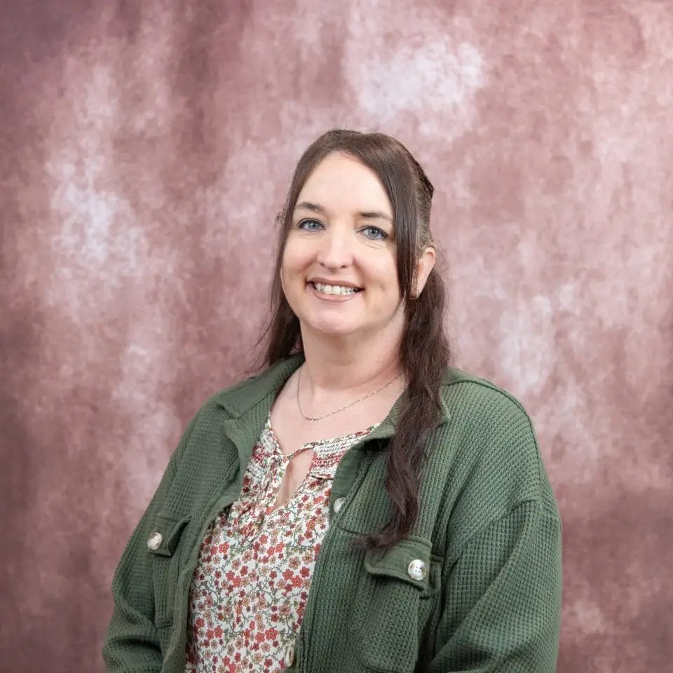 Smiling woman with long brown hair, blue eyes, wearing green jacket and floral top.